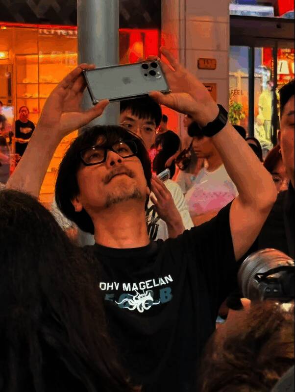 小岛秀夫在上海街头行走,身穿黑色外套,手持相机