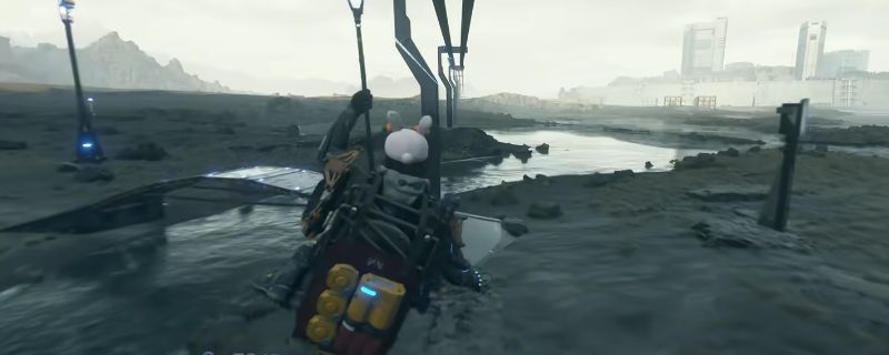 《死亡搁浅2》游戏中玩家手持改装后的消音狙击枪,在荒原环境中准备瞄准远处目标,枪械细节清晰可见