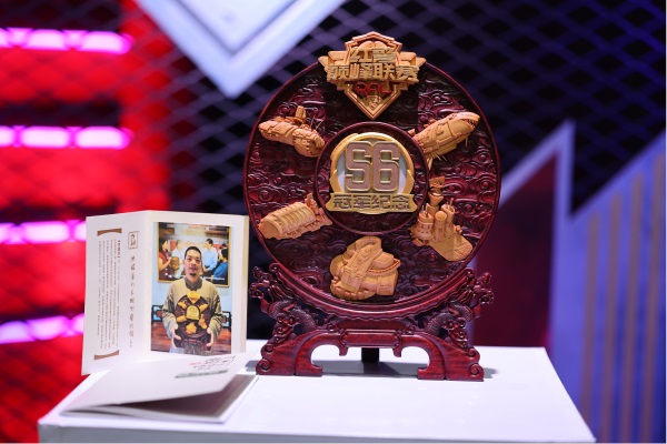 非遗大师洪建华正在雕刻红警主题奖杯，工作台上摆放精细木雕作品，细节精致