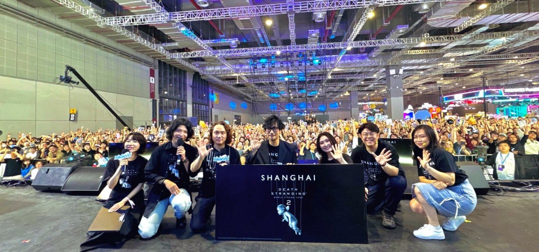 小岛秀夫与现场年轻玩家击掌互动特写，玩家手绘《死亡搁浅》主题T恤清晰可见，背景LED屏滚动播放中文配音预告片花