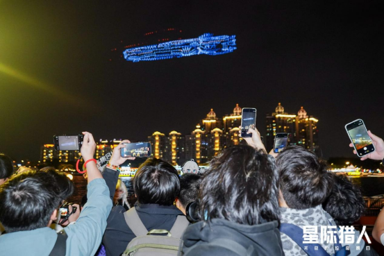 广州塔广场全景俯拍：数百名身着《星际猎人》主题服饰的玩家高举发光手幅，仰望夜空中的无人机舰队，人群与天幕上的‘盘古舰’形成天地呼应的沉浸式画面